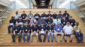 A group of about 28 people, mostly men, pose on wide wooden steps inside a modern building. Many wear matching dark shirts and hats, with some holding plaques or certificates.